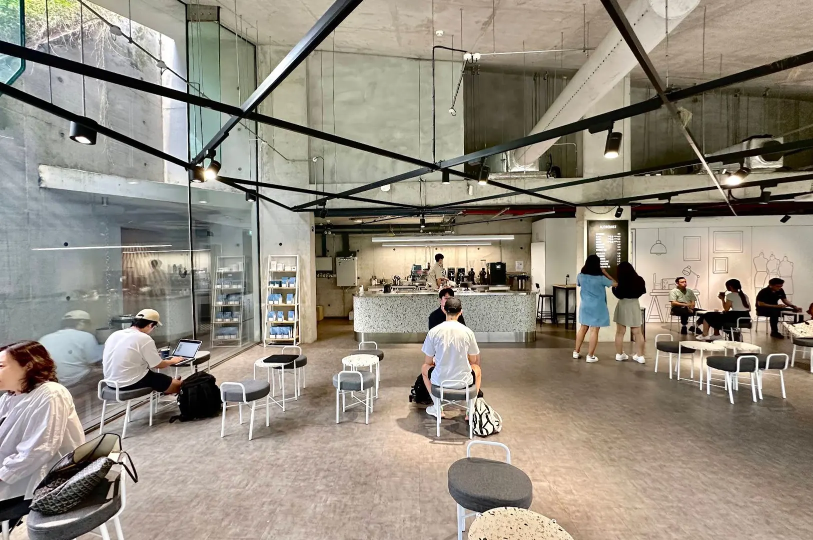 Patrons sit on scattered round stools and small tables throughout a spacious, industrial-style coffee shop with raw concrete walls and exposed black ceiling framework. In the background, baristas prepare beverages behind a curved terrazzo counter beneath bright track lighting.
