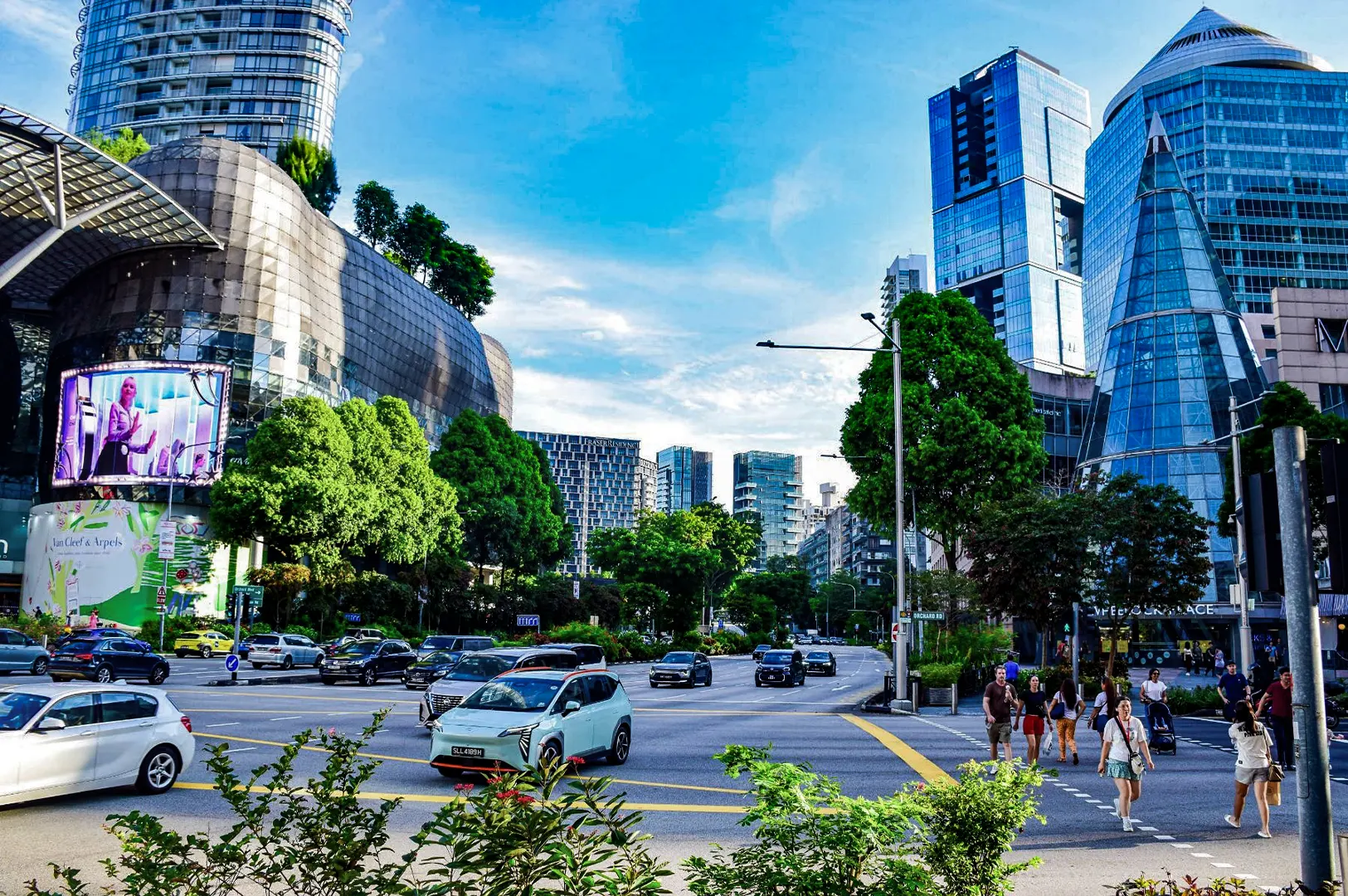 A vibrant street scene at Orchard Road in Singapore shows cars driving through a busy intersection lined with lush green trees and pedestrian crosswalks. Futuristic shopping malls and modern glass skyscrapers frame the street under a clear blue sky.