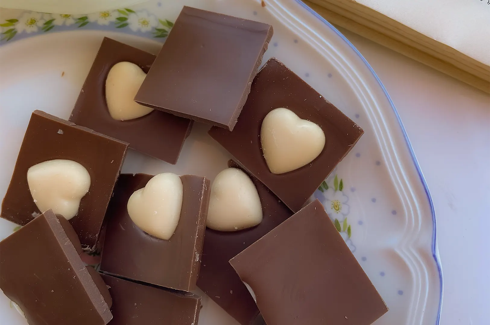 Square pieces of milk chocolate are arranged on a floral ceramic plate, with several squares embedded with white chocolate heart centers. An open book rests partially visible in the upper-right corner of the soft-lit surface.
