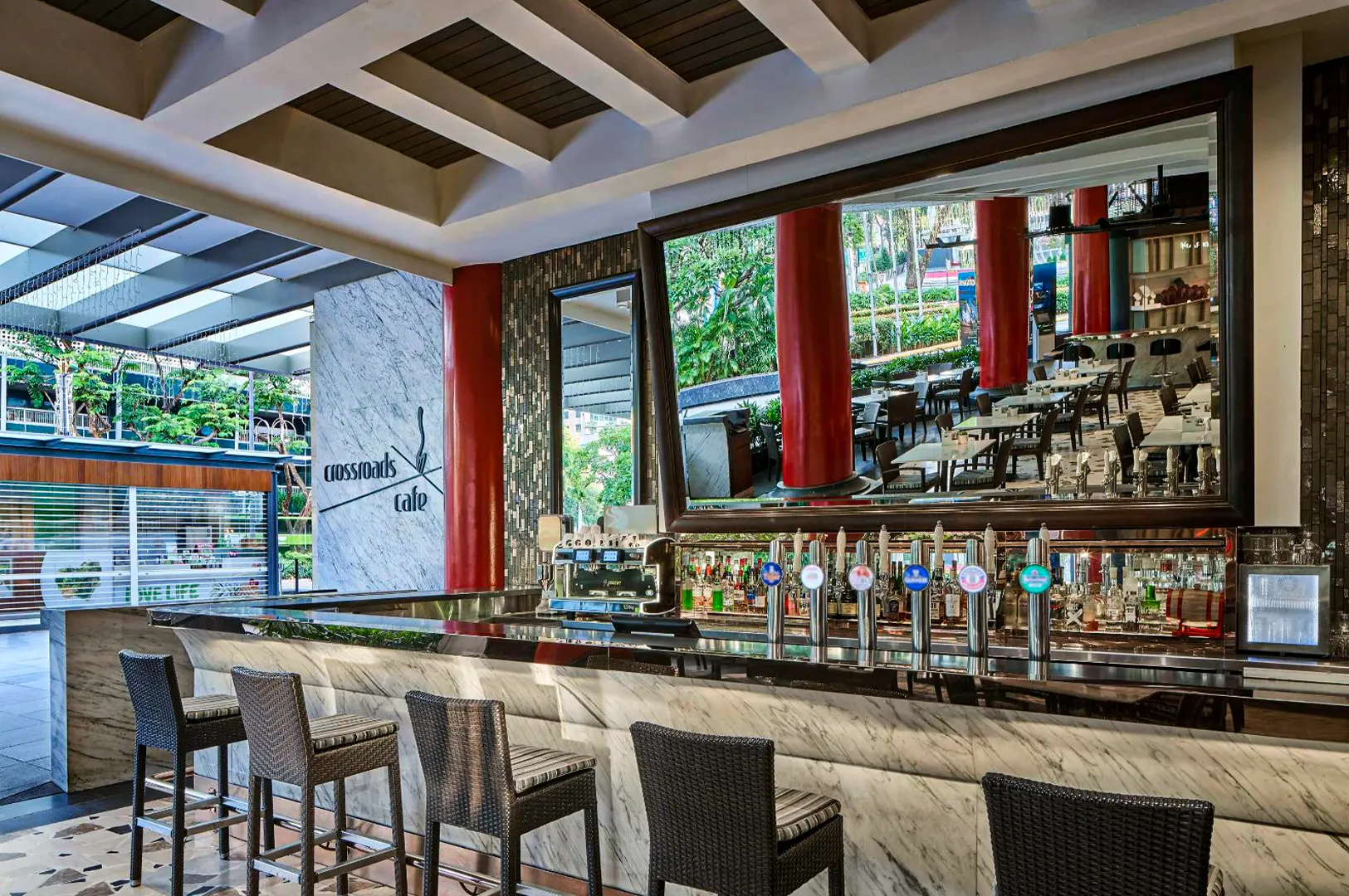 An open-air bar features a polished white marble counter lined with woven high-top barstools, chrome beer taps, and an espresso machine. A large rectangular mirror behind the counter reflects bold red pillars and the outdoor patio dining area of Crossroads Cafe.