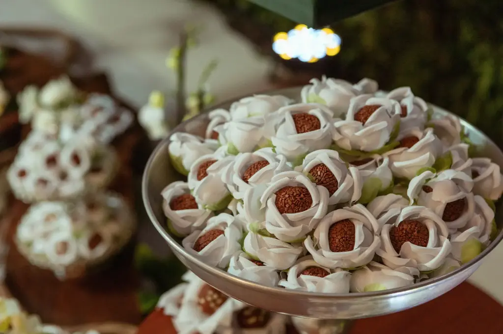 A silver pedestal platter holds chocolate brigadeiro candies wrapped in delicate white fabric flower cups designed to resemble open roses. Similar decorative dessert arrangements are softly blurred across the background table display.
