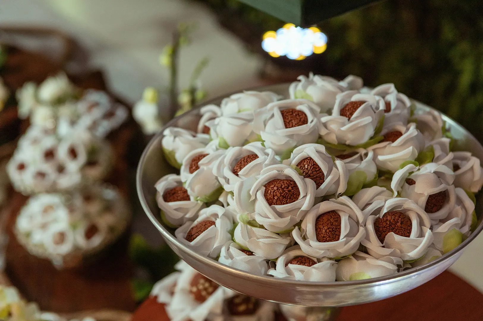 A silver pedestal platter holds chocolate brigadeiro candies wrapped in delicate white fabric flower cups designed to resemble open roses. Similar decorative dessert arrangements are softly blurred across the background table display.