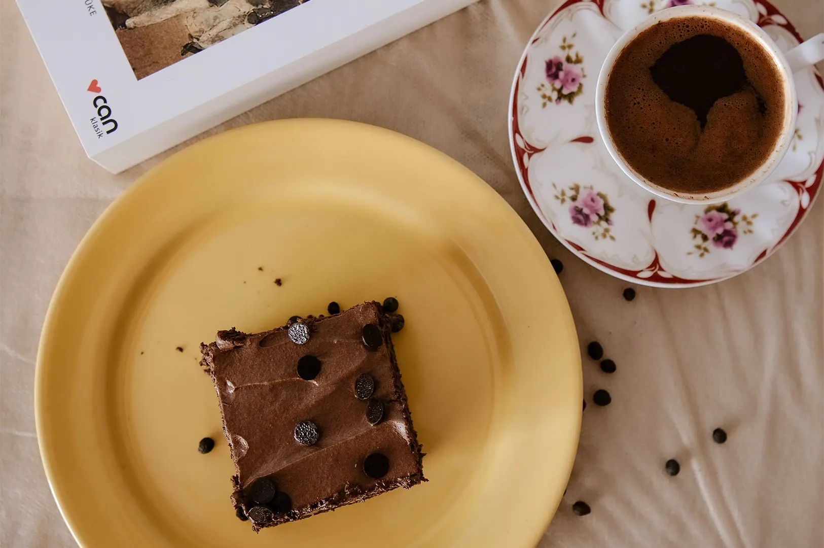 A top-down view shows a slice of rich chocolate cake topped with chocolate chips served on a pastel yellow ceramic plate. Beside it sits a floral teacup filled with dark coffee or espresso and a paperback book on a beige tablecloth sprinkled with coffee beans.
