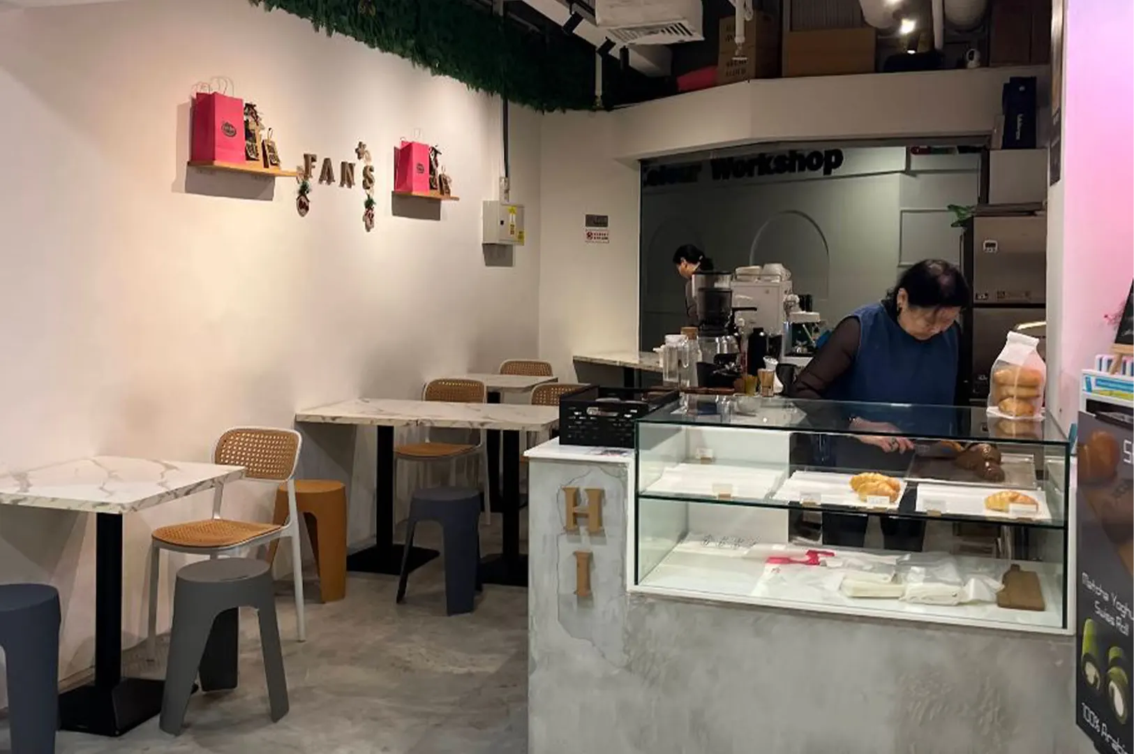 A cozy café interior features small white marble tables paired with woven-back chairs and plastic stools along a plain wall decorated with pink shopping bags and lettering spelling "FAN'S". In the foreground, a staff member works behind a glass pastry display counter filled with fresh baked goods.