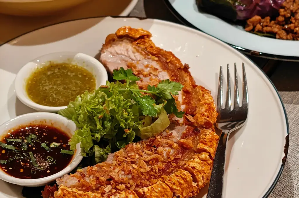 Crispy, golden-brown roasted pork belly slices with crackling skin are served on a white plate alongside fresh leafy greens and cilantro. Two small ramekins containing green chili sauce and a dark dipping sauce sit on the plate with a silver fork.