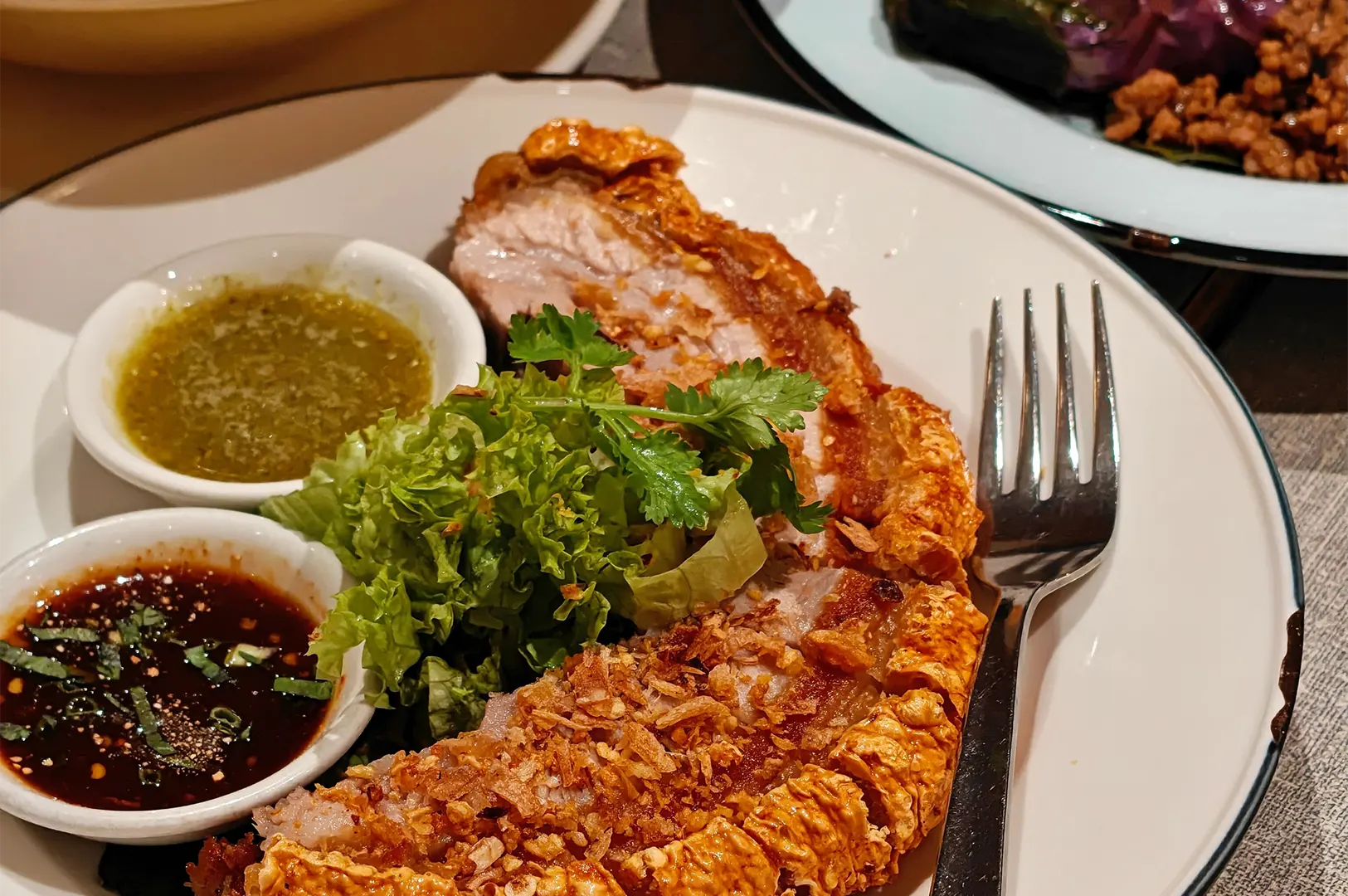 Crispy, golden-brown roasted pork belly slices with crackling skin are served on a white plate alongside fresh leafy greens and cilantro. Two small ramekins containing green chili sauce and a dark dipping sauce sit on the plate with a silver fork.