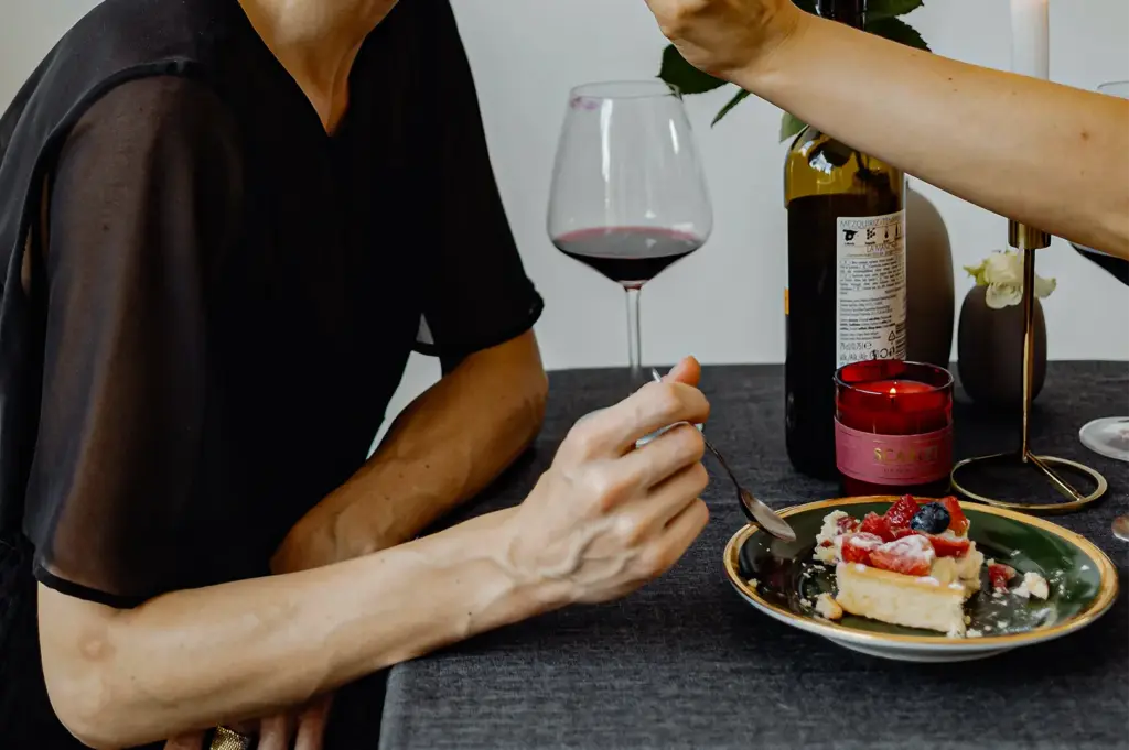 A close-up shot captures a person seated at a dark-clothed table eating a slice of cheesecake topped with fresh berries. In the background, a bottle of wine, a wine glass, a small lit candle, and another reaching hand set an intimate dining scene.