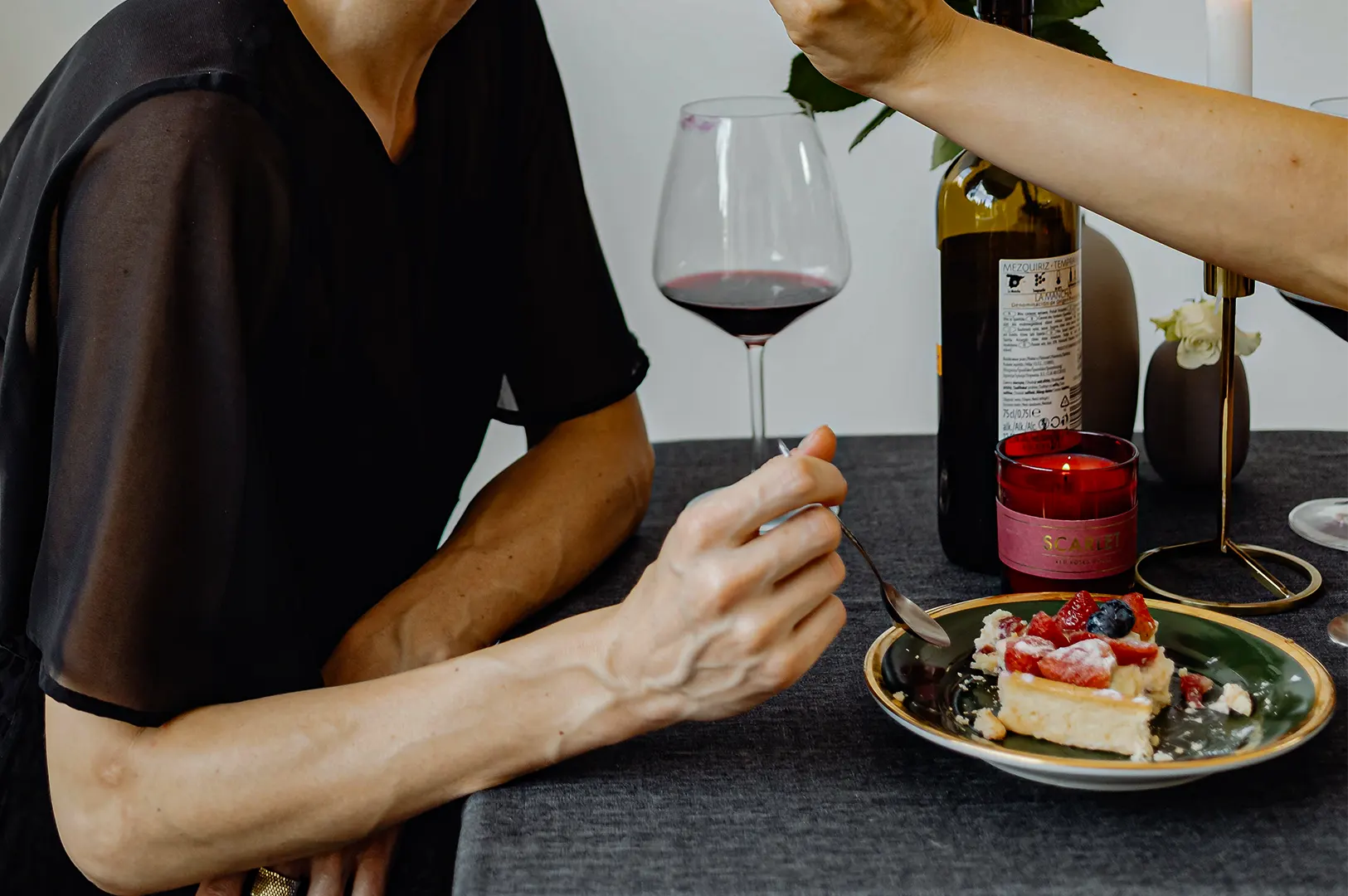 A close-up shot captures a person seated at a dark-clothed table eating a slice of cheesecake topped with fresh berries. In the background, a bottle of wine, a wine glass, a small lit candle, and another reaching hand set an intimate dining scene.