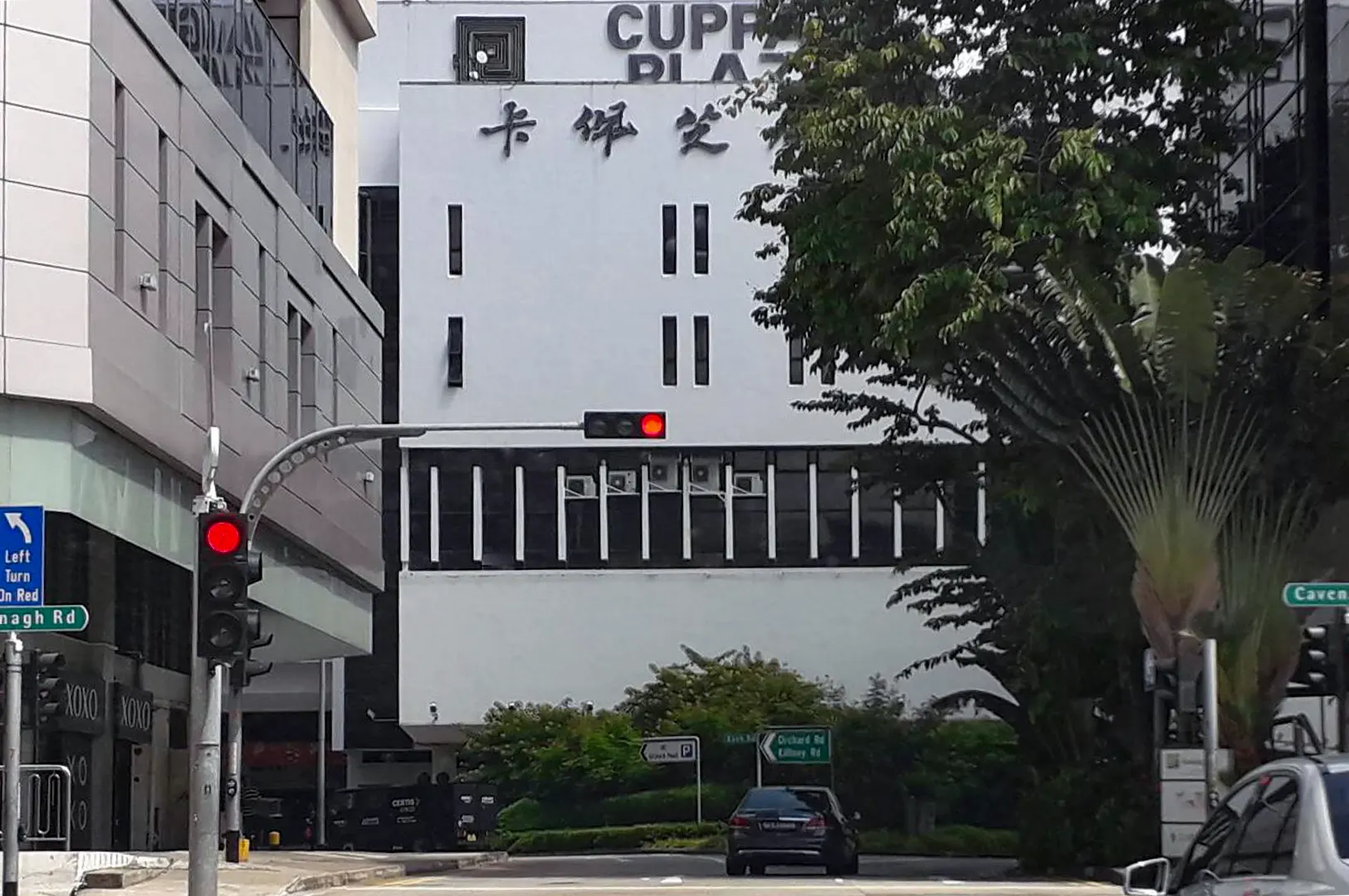 An exterior street view shows the facade of Cuppage Plaza with English and Chinese signage mounted on its white building face. A dark car sits at a red traffic light on the road in the foreground, flanked by street signs and lush green trees.