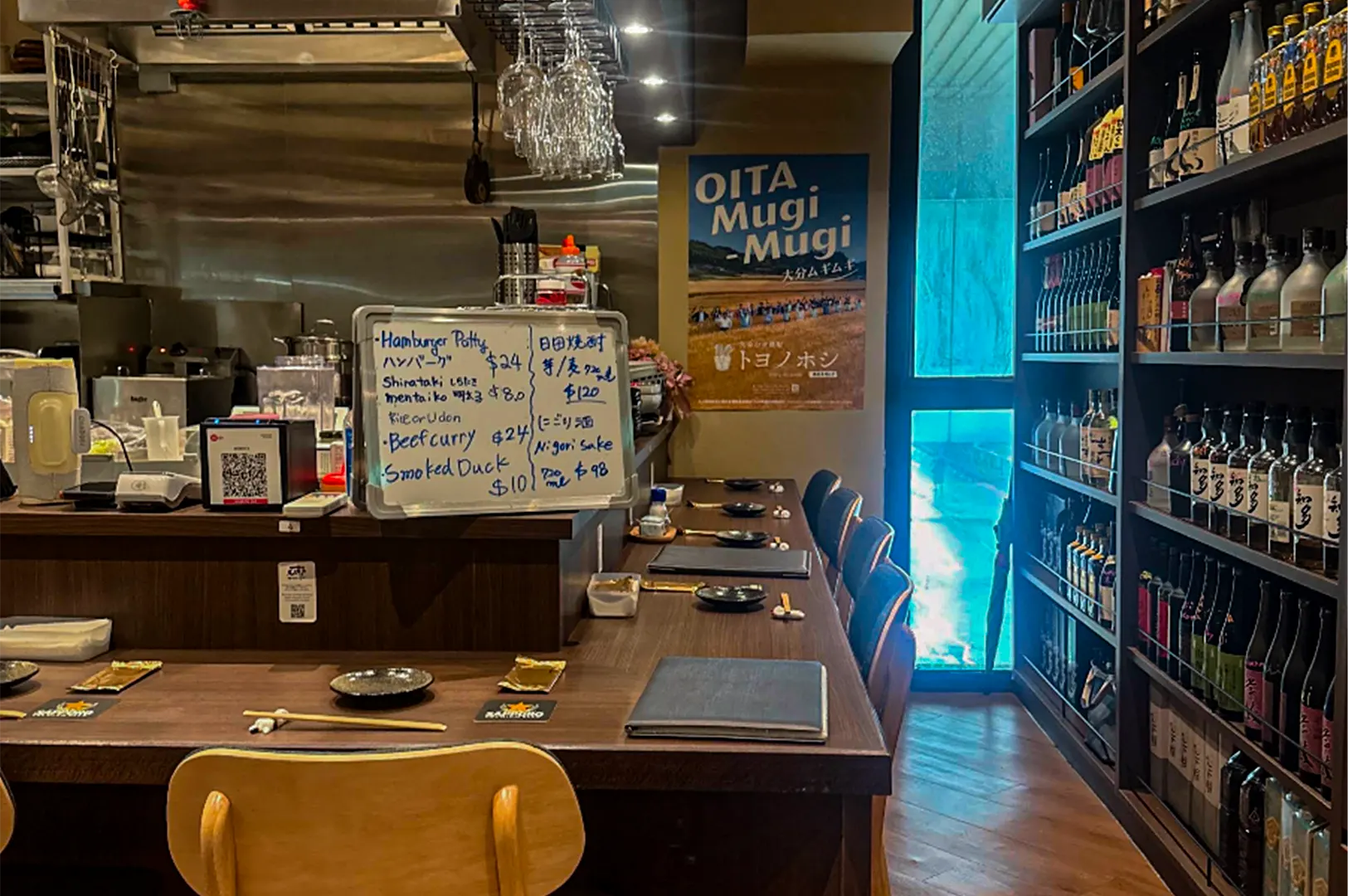 An L-shaped wooden counter bar is set with place settings, menus, and a handwritten whiteboard listing daily food and drink specials. Shelves filled with bottled sake and spirits line the right wall, while wine glasses hang above the open kitchen area.