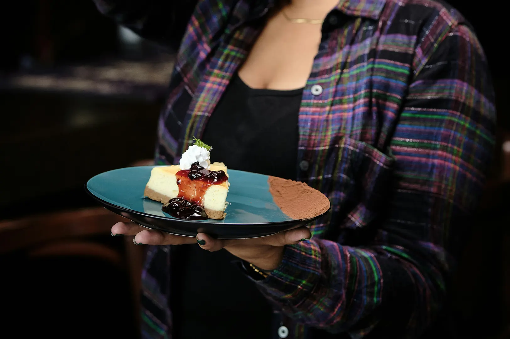 A person wearing a colorful plaid shirt holds out a teal plate featuring a slice of blueberry cheesecake. The dessert is topped with whipped cream, fresh herb garnish, and berry compote, with cocoa powder dusted along one edge of the plate.