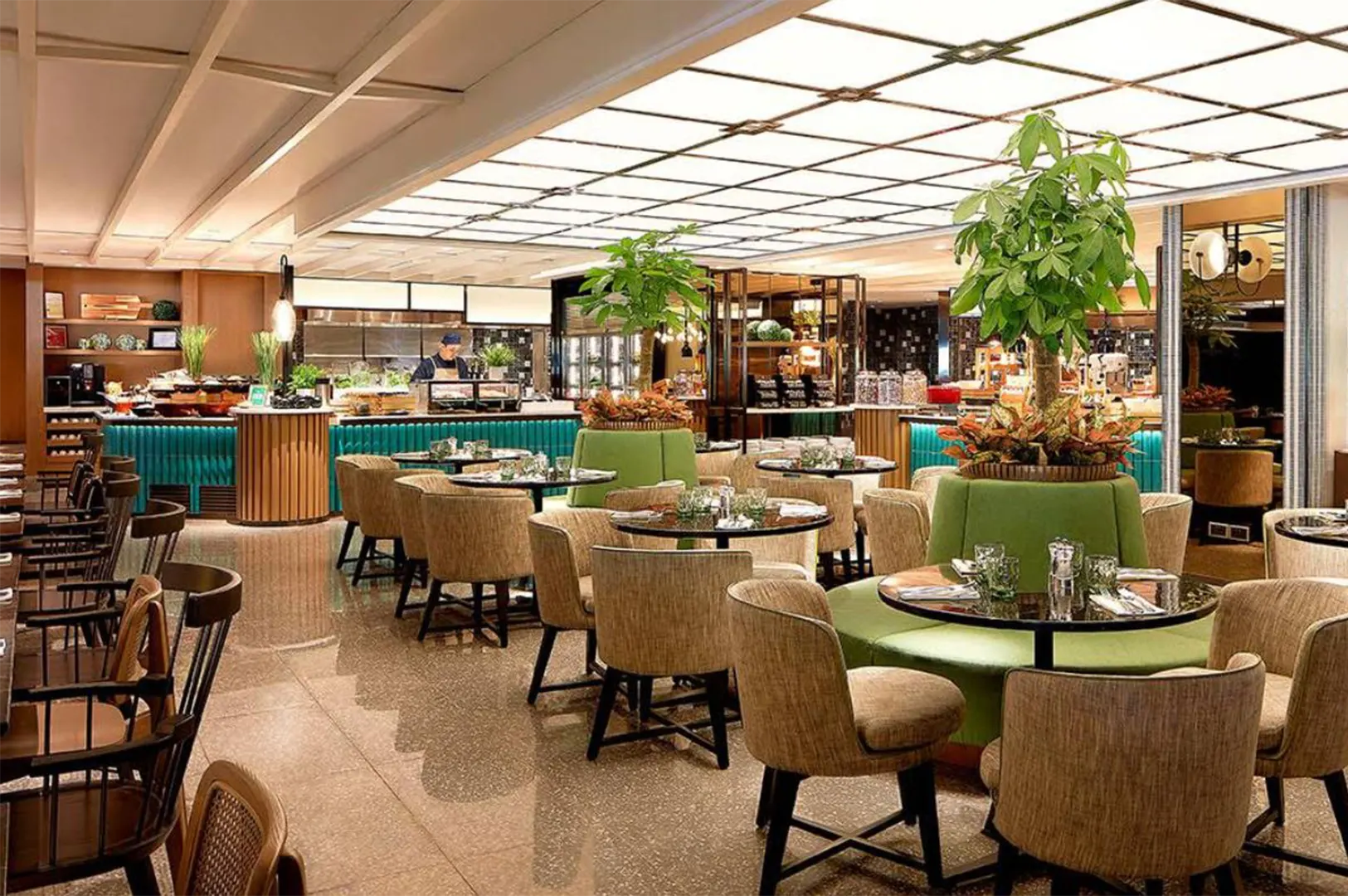 A bright, spacious buffet restaurant featuring plush beige chairs, polished tiled floors, and vibrant green circular booths centered around potted indoor trees. In the background, modern food display counters and live cooking stations stand under a glowing, backlit ceiling grid.