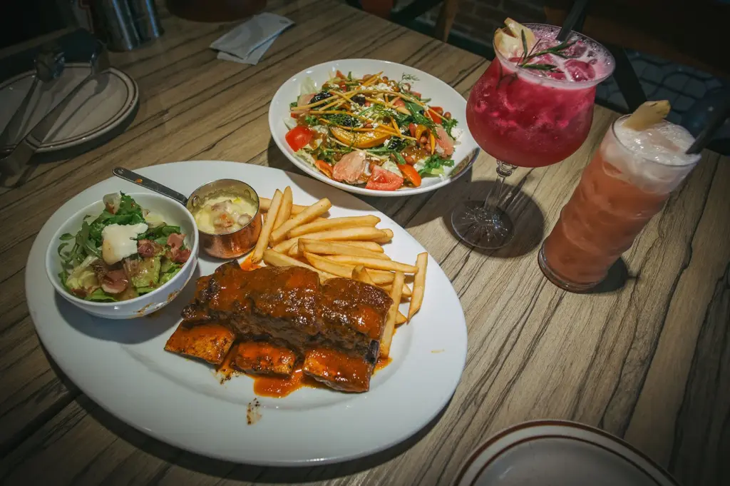 A dinner spread served on a wooden table features a main platter with sauce-covered ribs, French fries, a small green salad, and a ramekin of cheese dip. In the background, a large bowl of fresh garden salad rests next to two colorful iced cocktails garnished with fruit and rosemary.
