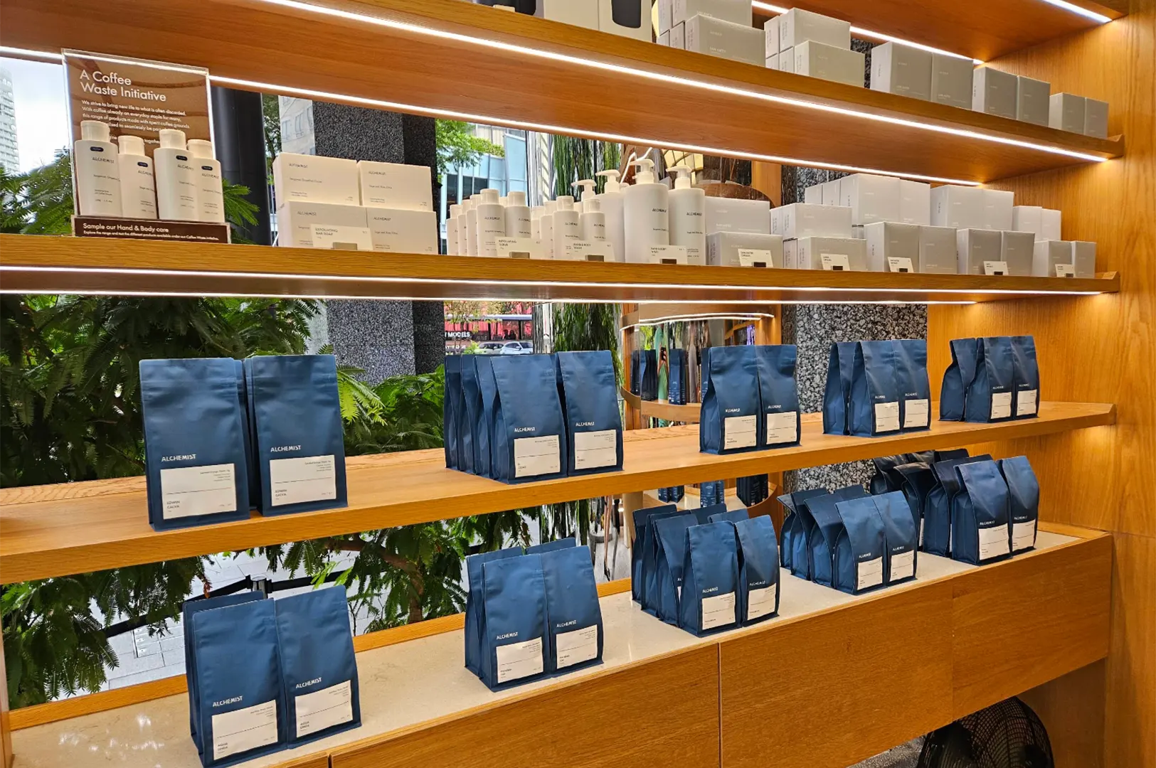Neatly illuminated wooden retail shelves display rows of packaged coffee products and skincare items. The lower tiers are filled with blue Alchemist coffee bags, while the upper shelves feature white boxes and pump bottles alongside a framed informational sign.