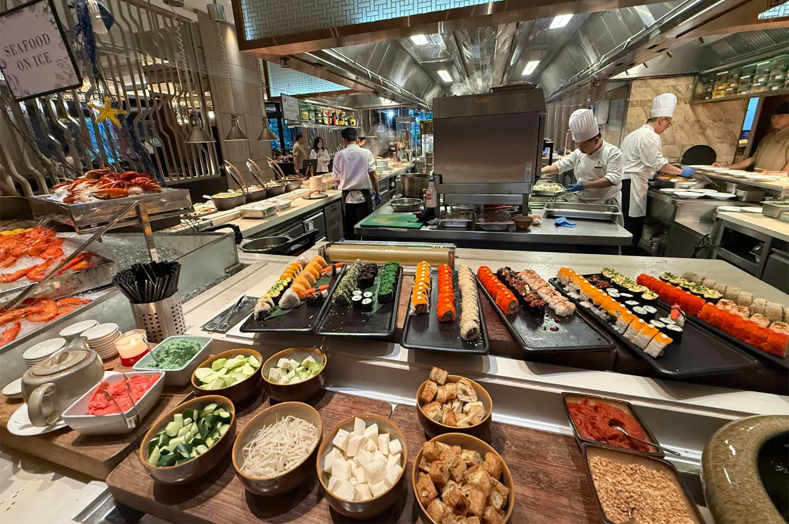 Chefs in tall white hats prepare food behind a bustling buffet line stocked with colorful sushi rolls, fresh seafood on ice, and bowls of soup toppings. The wide display features neatly organized rows of maki rolls, chilled prawns, crabs, cut vegetables, and condiments.