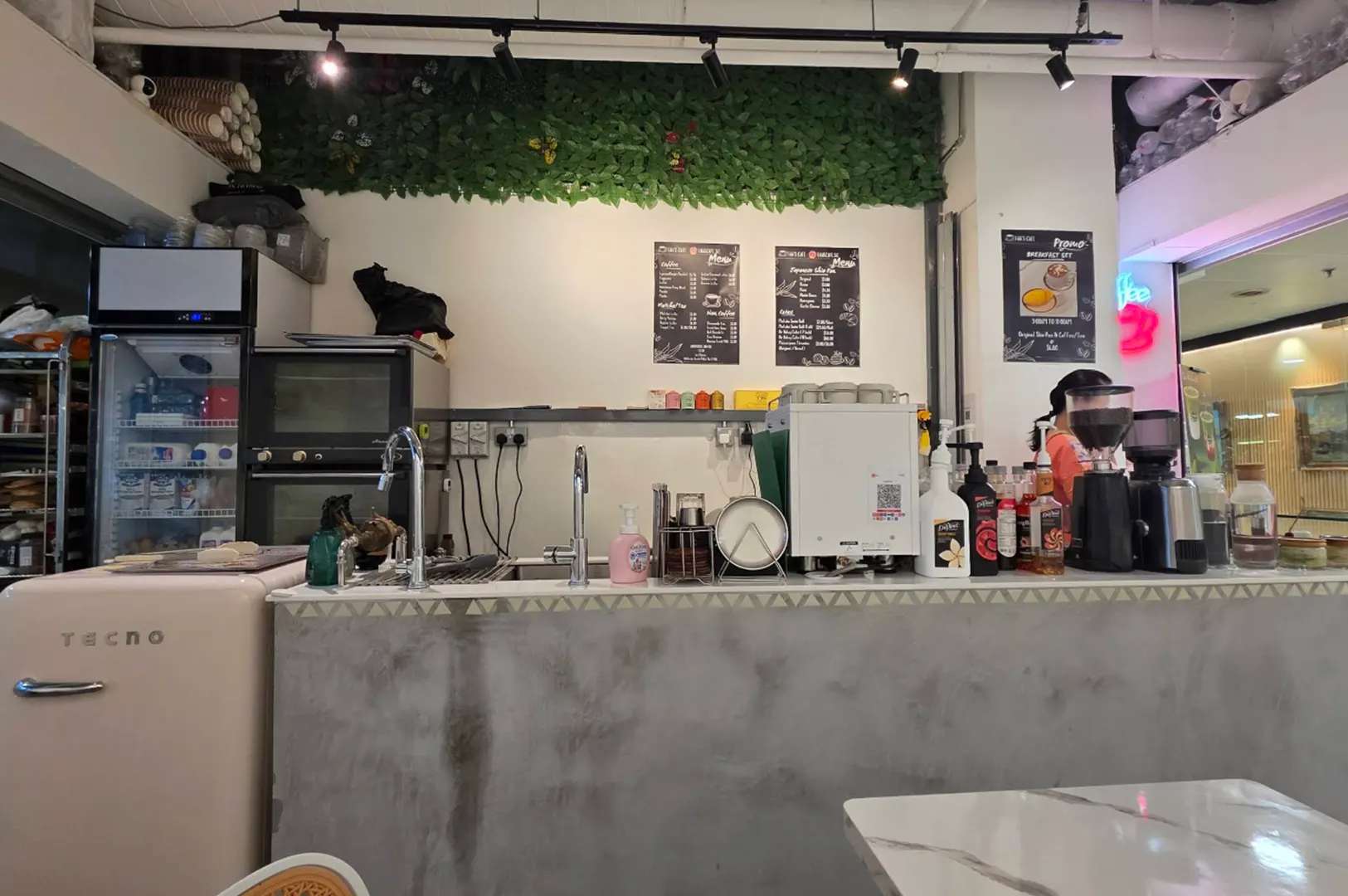 A compact coffee shop service counter features an espresso machine, coffee grinders, syrup bottles, and sinks set against a concrete-finish bar. Black chalkboards displaying drink menus hang on the wall below overhead track lights and a strip of decorative artificial greenery.
