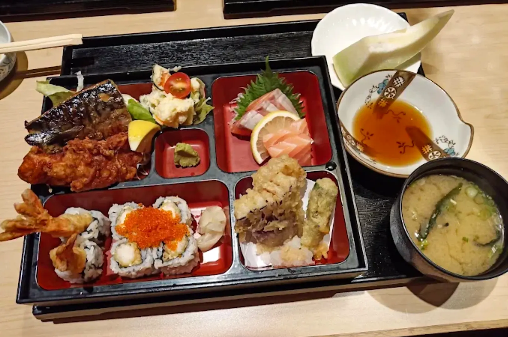 A traditional Japanese bento box is neatly arranged with grilled fish, chicken karaage, fresh sashimi slices, sushi rolls topped with tobiko, vegetable tempura, and potato salad. The set meal is served on a black tray alongside a bowl of miso soup, dipping sauce, and a fresh melon wedge.