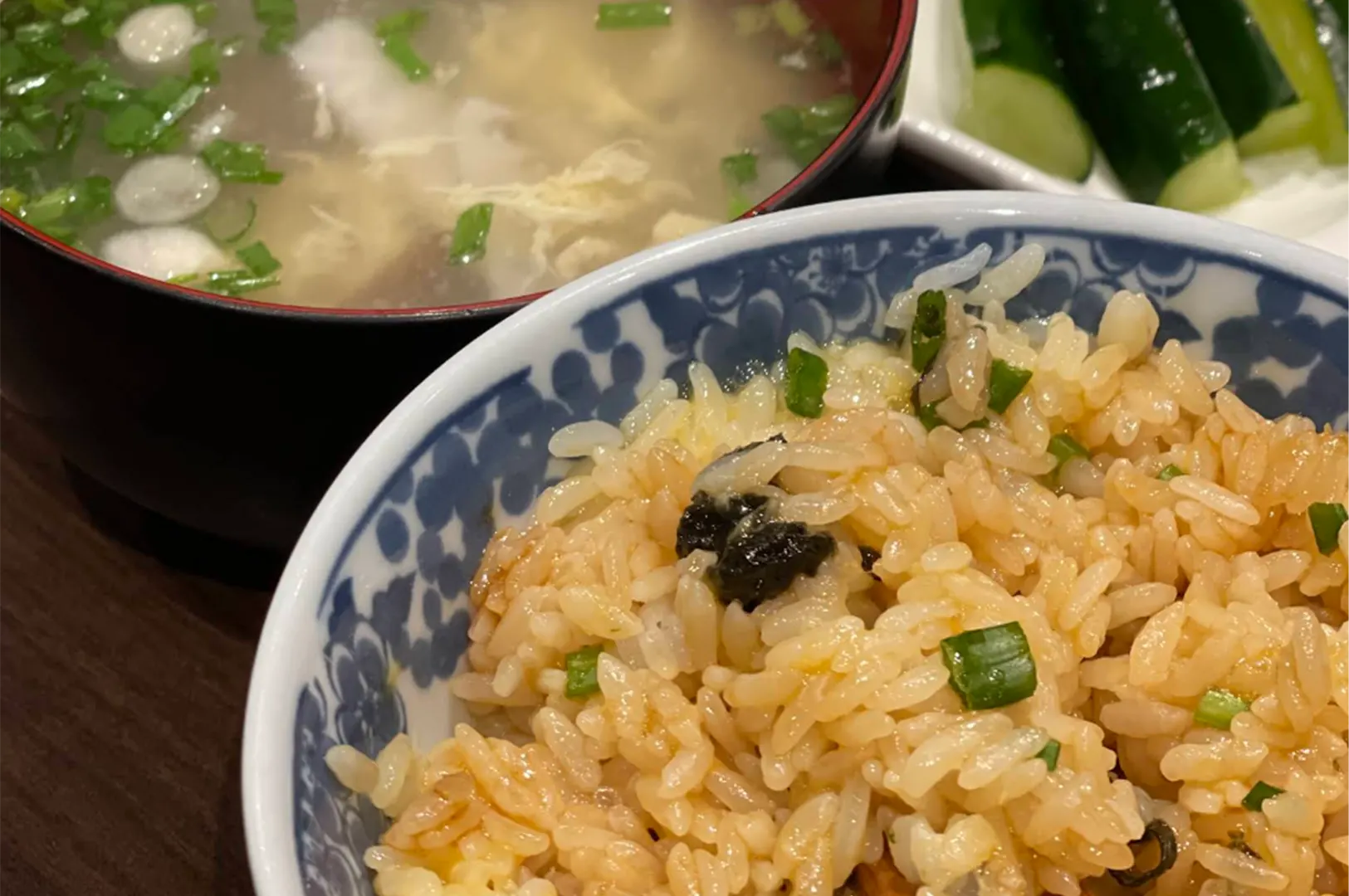 A blue-and-white patterned ceramic bowl holds seasoned Japanese fried rice mixed with chopped scallions and seaweed. Beside it, a bowl of clear broth with egg drops and green onions rests next to a side plate of sliced cucumbers.