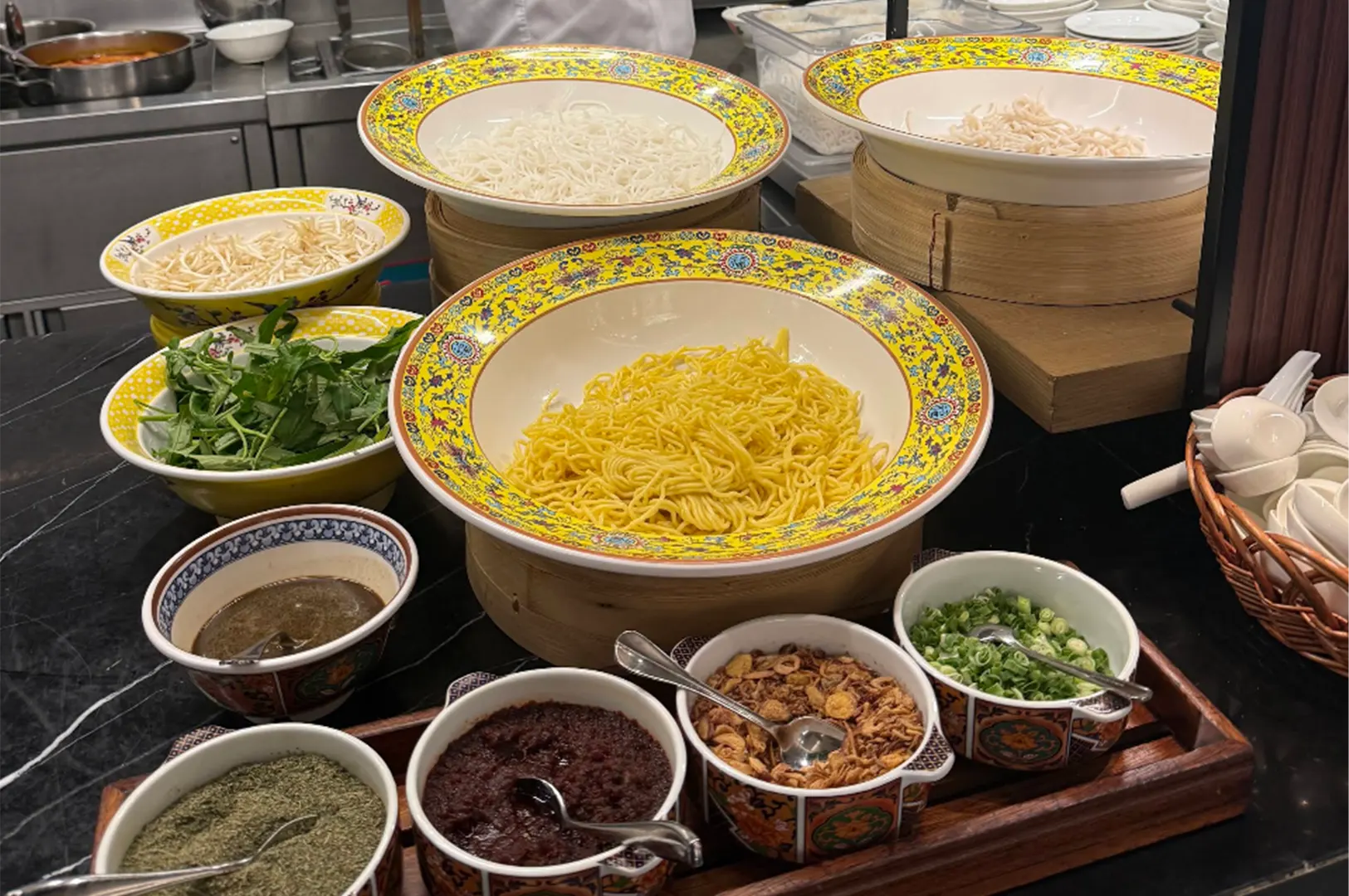 A noodle station at a buffet display features yellow egg noodles, white rice noodles, bean sprouts, and fresh greens in ornate yellow bowls rested on bamboo steamers. In the foreground, a wooden tray holds small bowls of condiments and toppings, including chili paste, crispy fried shallots, chopped scallions, and herbs.