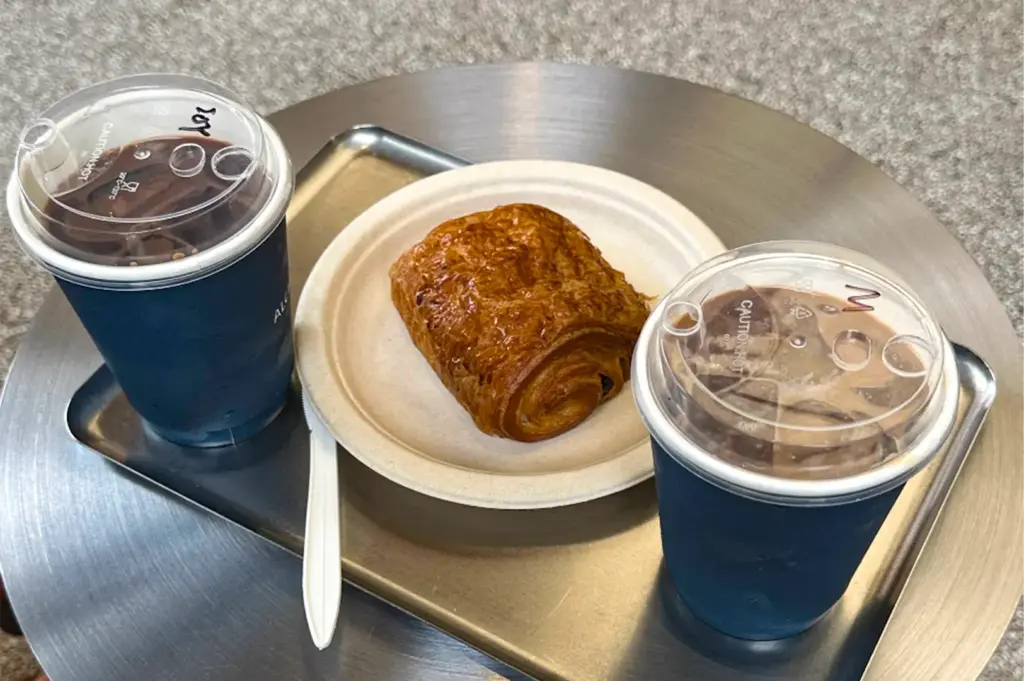 Two dark blue paper cups with clear plastic sip lids sit on a metallic tray alongside a golden pain au chocolat on a white paper plate. The iced beverage order is placed on a round metal tabletop, accompanied by a plastic knife.