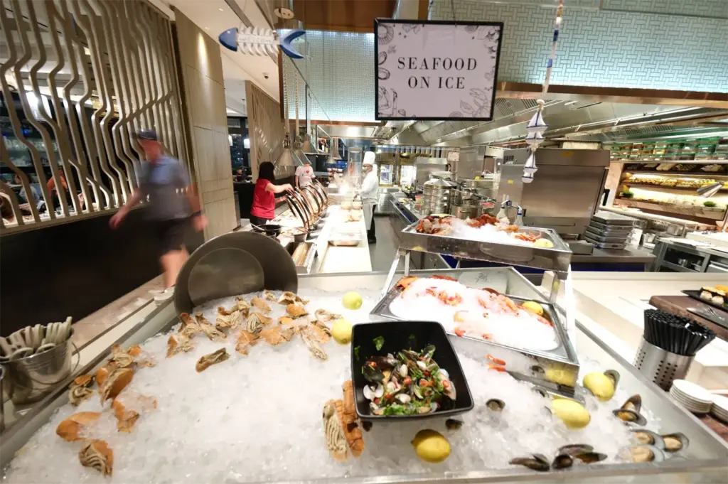A "Seafood On Ice" buffet station features a large bed of crushed ice laden with slipper lobsters, mussels, prawns, lemons, and a bowl of marinated shellfish. Diners walk past hot food chafing dishes and a chef at work in the brightly lit background.