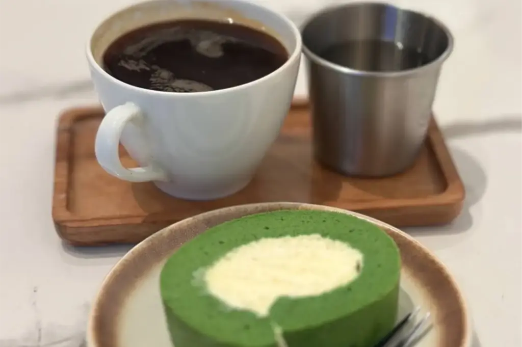 A slice of vibrant green matcha swiss roll with a light cream filling sits in the foreground on a ceramic plate. In the background, a white ceramic mug of black coffee and a small stainless steel cup rest on a rectangular wooden tray.