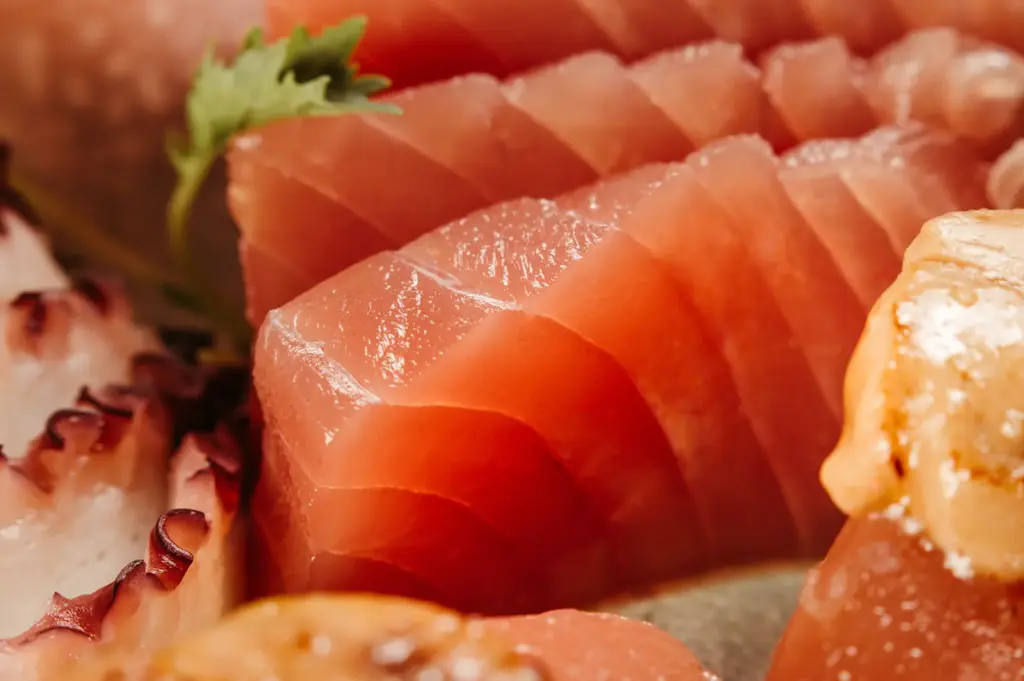 A detailed close-up shot highlights freshly sliced, thick-cut raw tuna sashimi showing fine grain and texture. Slices of octopus tentacle with suction cups and other pieces of seafood sit alongside the tuna in soft focus.