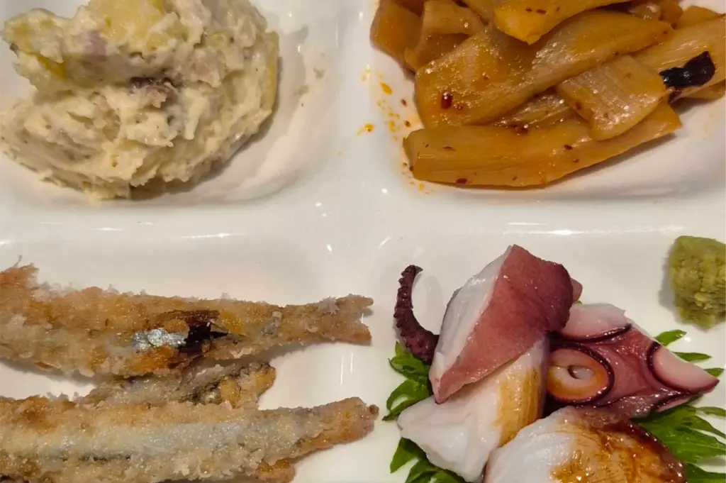 A four-compartment white serving platter presents an assortment of Japanese side dishes, including creamy potato salad, seasoned bamboo shoots (menma), crispy fried whole fish, and sliced octopus sashimi on a green shiso leaf with a dab of wasabi.