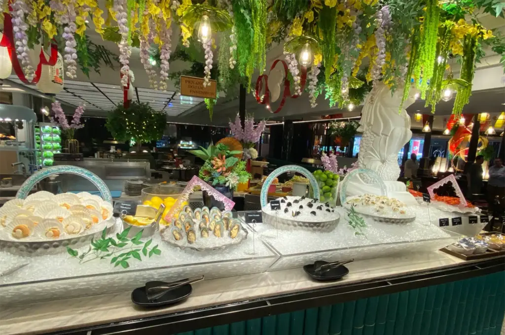 A lavish seafood buffet display features fresh scallops, green mussels, black mussels, and shellfish presented over crushed ice under hanging yellow and purple floral garlands. A white Merlion statue sits at the end of the counter next to additional seafood selections.