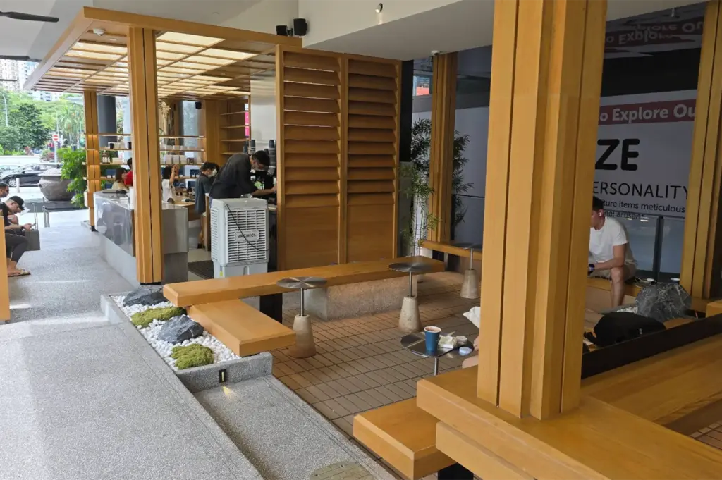 A modern open-air coffee kiosk constructed with warm wooden pillars and slatted paneling features bench seating and small side tables. Baristas work behind a sleek service counter on the left, while patrons relax in the surrounding shaded seating area.