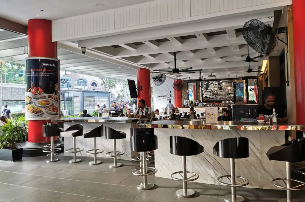 Patrons sit at a marble-topped bar with sleek black swivel stools in an open-air cafe setting framed by prominent red pillars. Modern ceiling fans hang overhead, and promotional posters advertise dining options alongside views of the surrounding city street.