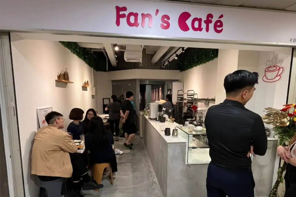 The storefront view of Fan's Café displays a bold pink logo above the open entrance, revealing a busy, narrow interior filled with customers seated along the left wall. A long service counter stretches along the right side toward the back, with staff working behind the bar and a man standing in the right foreground.