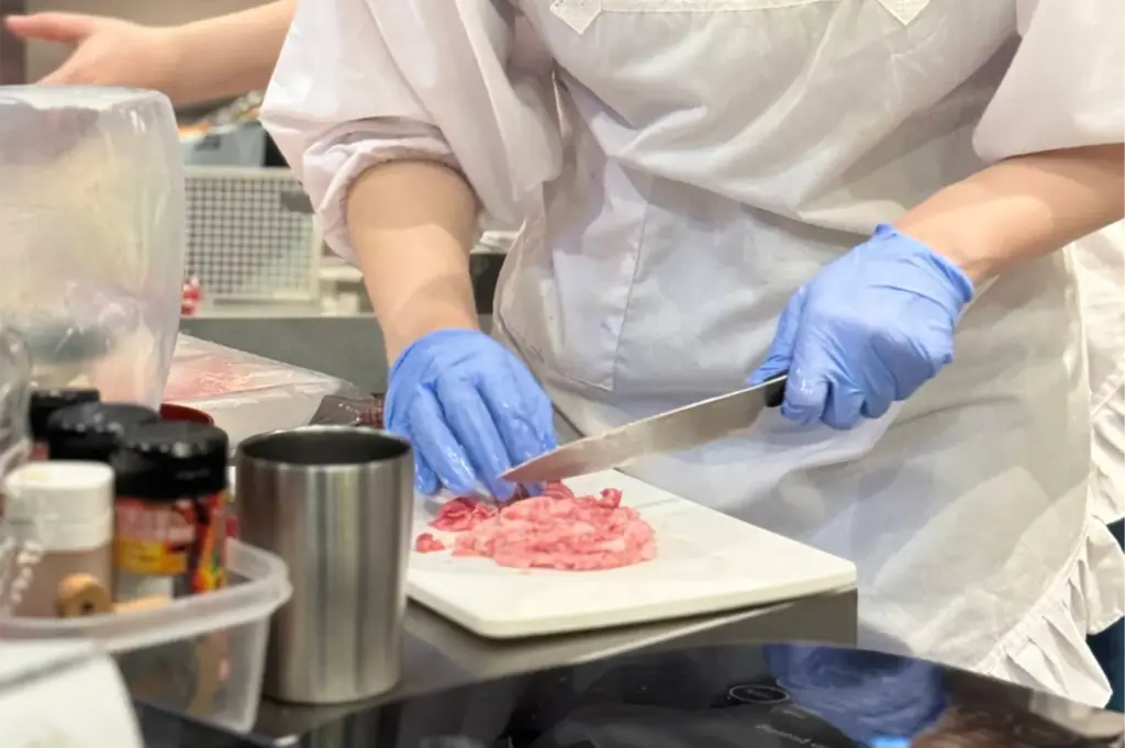 A cook wearing blue disposable gloves and a white apron carefully slices thin pieces of raw beef on a white cutting board. Seasoning jars, a stainless steel cup, and other kitchen prep tools sit nearby on the counter.