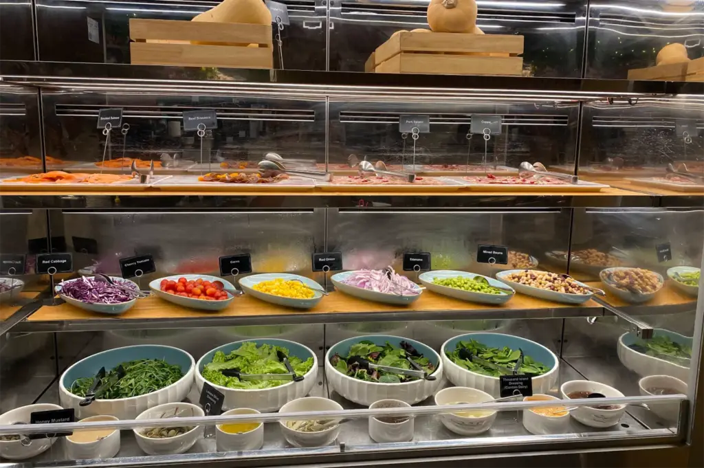 A refrigerated glass display case is stocked with cold cuts and cured meats on the top shelf, colorful salad toppings like corn and cherry tomatoes on the middle shelf, and fresh leafy greens with small bowls of dressings along the bottom. Whole butternut squashes rest in wooden crates above the case.