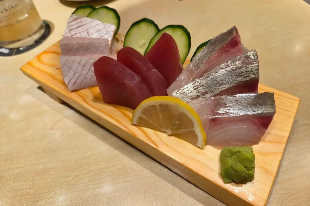 A wooden platter displays an array of fresh raw sashimi, including thick-cut tuna, pale pink whitefish, and silver-skinned fish slices. The presentation is garnished with thin cucumber rounds, a lemon wedge, and a dollop of green wasabi paste on the side.