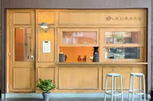 A minimalist light-wood storefront for "Kurasu" features clean paneling, a glass-paneled entry door, and a service counter equipped with pour-over coffee drippers. Two light grey bar stools stand outside beside a potted green plant, giving the cafe a warm, contemporary aesthetic.