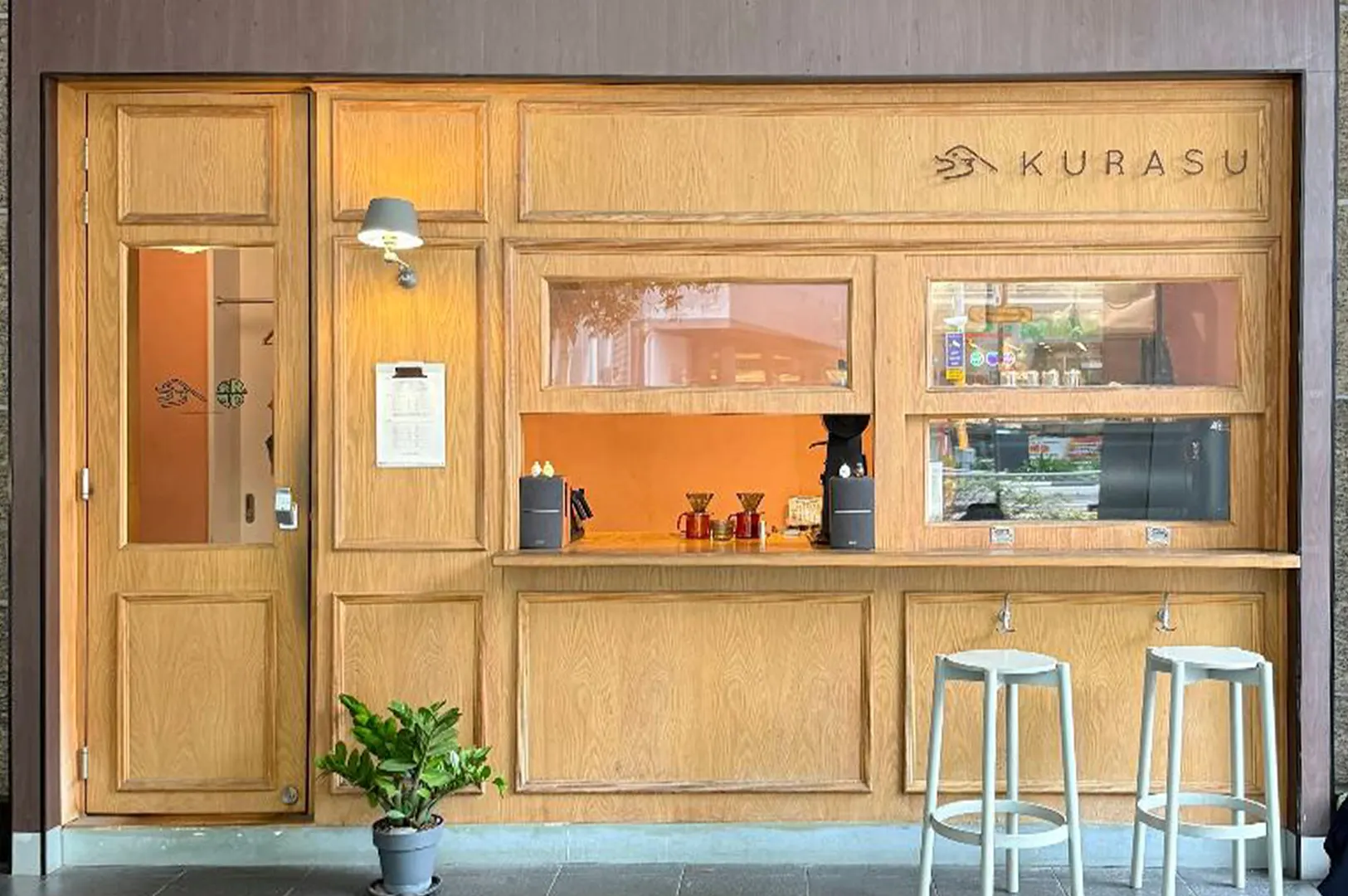 A minimalist light-wood storefront for "Kurasu" features clean paneling, a glass-paneled entry door, and a service counter equipped with pour-over coffee drippers. Two light grey bar stools stand outside beside a potted green plant, giving the cafe a warm, contemporary aesthetic.