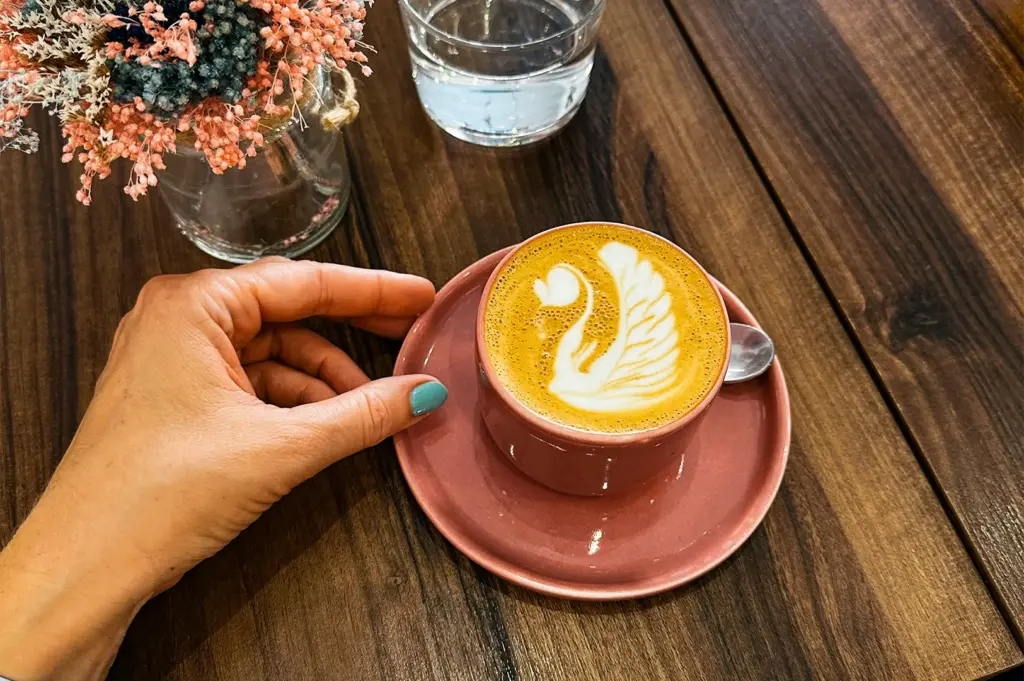 A hand with turquoise-painted nails reaches toward a pink ceramic cup of latte featuring intricate swan latte art on its saucer. Beside the coffee, a glass of water and a small vase of colorful dried flowers rest on the dark wooden table, capturing a quiet cafe moment.