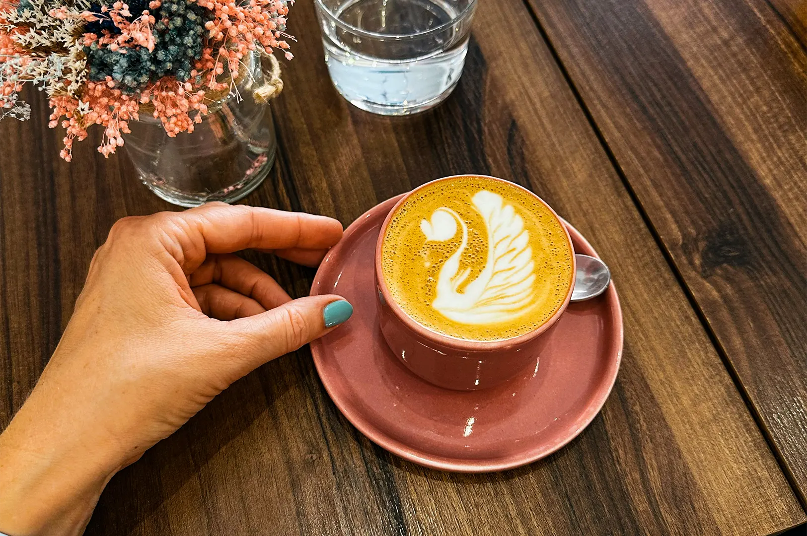 A hand with turquoise-painted nails reaches toward a pink ceramic cup of latte featuring intricate swan latte art on its saucer. Beside the coffee, a glass of water and a small vase of colorful dried flowers rest on the dark wooden table, capturing a quiet cafe moment.
