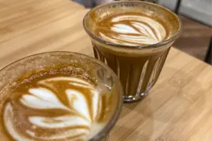 Two small, clear fluted glasses filled with espresso-based coffees sit side by side on a light wooden table surface. Both drinks are topped with delicate white steamed milk latte art designs swirled into the rich brown crema.
