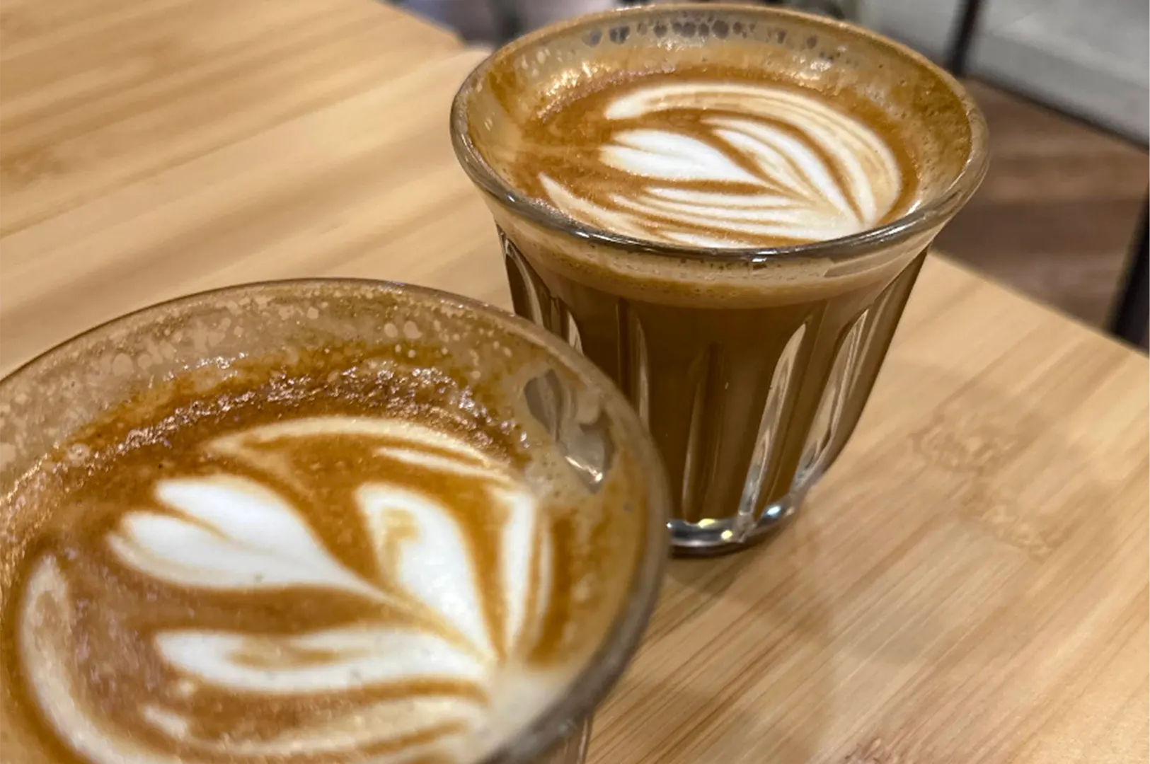 Two small, clear fluted glasses filled with espresso-based coffees sit side by side on a light wooden table surface. Both drinks are topped with delicate white steamed milk latte art designs swirled into the rich brown crema.