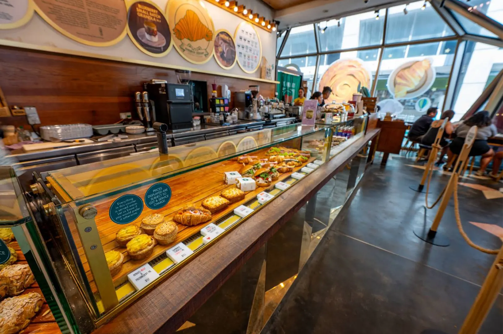 A long glass display counter inside a bustling modern bakery showcases a wide variety of fresh pastries, savory tarts, and filled croissants. Warm wooden paneling, circular wall signage, and large geometric windows line the interior where patrons sit in the background.