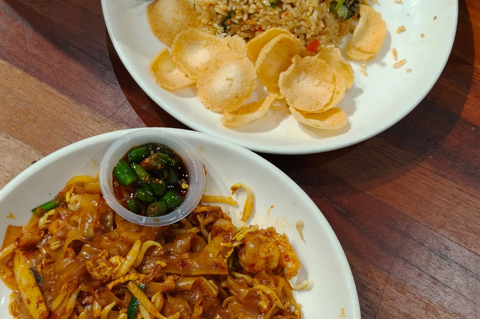 Two white plates of Southeast Asian cuisine are set on a dark wood table, with stir-fried flat rice noodles accompanied by a small plastic cup of sliced green chilies in soy sauce in the foreground. Beside it, a plate holds fried rice served with a generous topping of crispy golden crackers.