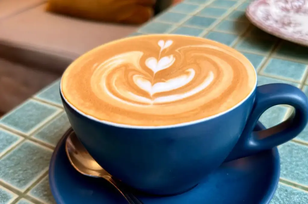 A matte blue ceramic cup filled with a hot latte rests on a matching saucer alongside a small spoon. The steamed milk foam features intricate latte art depicting stacked heart patterns, sitting atop a green tiled cafe tabletop.