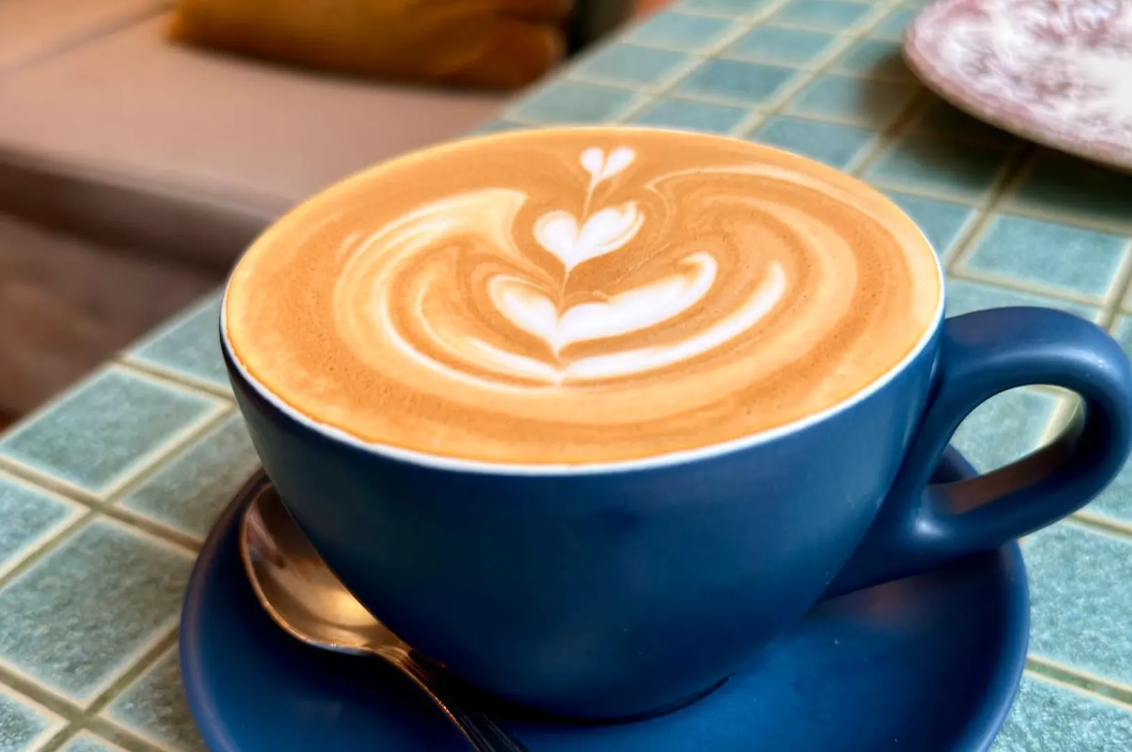 A matte blue ceramic cup filled with a hot latte rests on a matching saucer alongside a small spoon. The steamed milk foam features intricate latte art depicting stacked heart patterns, sitting atop a green tiled cafe tabletop.