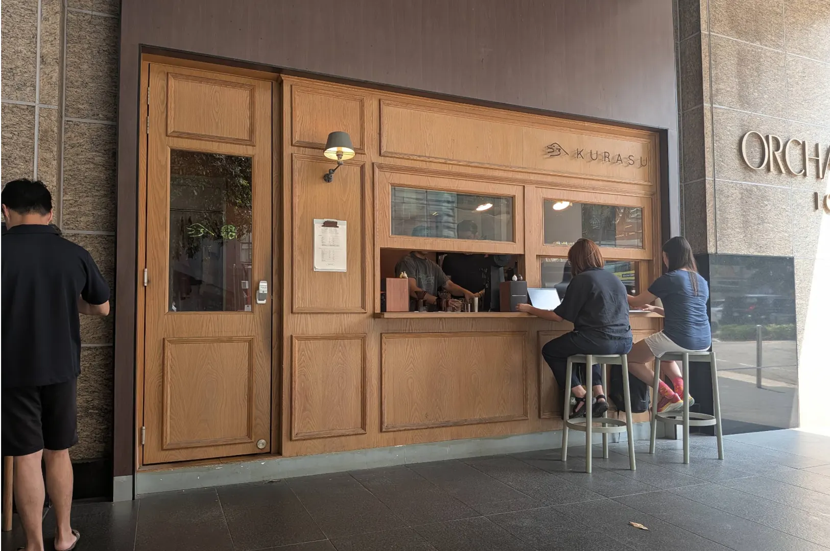 Two customers sit on high stools at the wooden outdoor counter of Kurasu, working on a laptop while barista staff prepare coffee inside the window service hatch. The Japanese-style coffee stand is set against a modern building façade with warm timber panelling and soft exterior lighting.