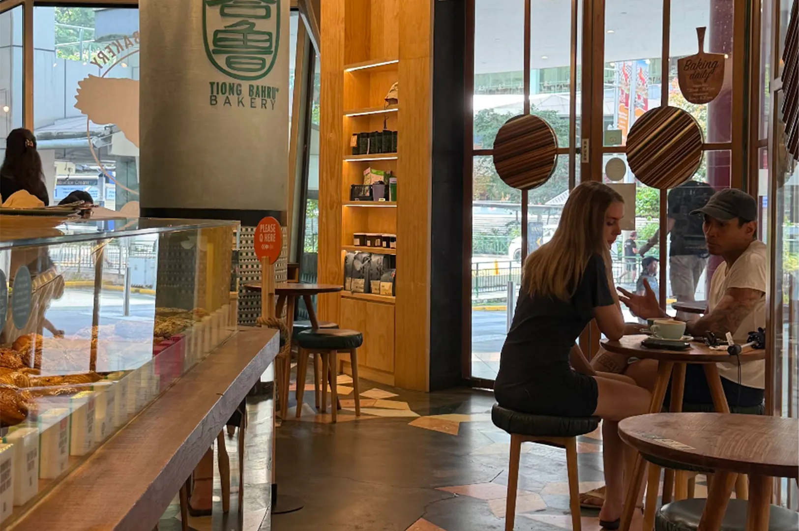 A couple sits at a small wooden table conversing over coffee inside a bright Tiong Bahru Bakery cafe. In the foreground, a glass pastry display case and wooden counters frame the warm, modern interior filled with light wood furnishings and large glass windows.