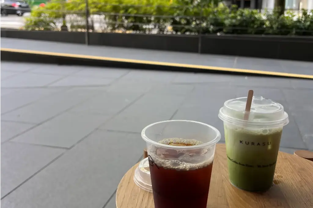 Two iced beverages—a dark iced filter coffee and a creamy iced green matcha latte in a branded Kurasu takeaway cup—rest on a light wooden table outdoor. In the background, a tiled urban walkway with lush greenery borders the sleek, open-air seating space.