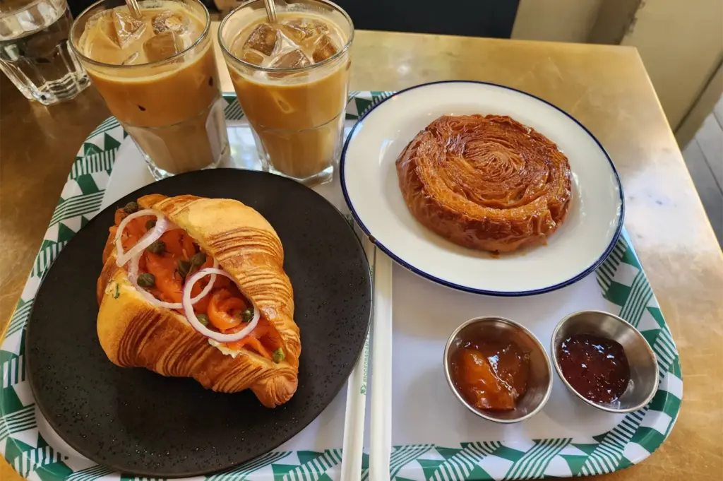 A patterned tray holds a delicious cafe spread featuring a smoked salmon croissant sandwich with red onion and capers alongside a golden kouign-amann pastry on a white plate. Two glasses of iced lattes and small ramekins of jam complete the table setup.