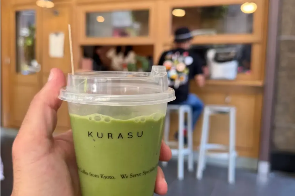 A close-up view shows a hand holding a clear, plastic cup filled with vibrant green iced matcha latte, clearly printed with the Kurasu logo. In the softly blurred background, a customer sits on a white stool facing the warm, wooden service window of the coffee stand.